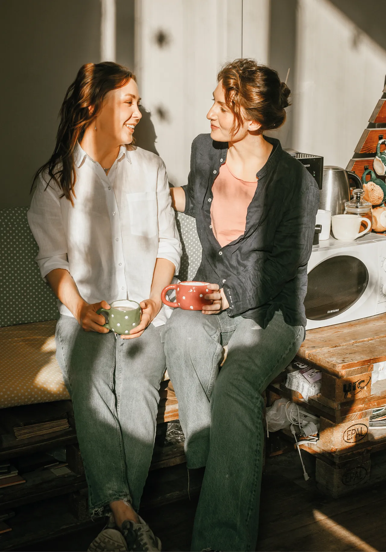 Zwei Frauen sitzen entspannt mit Tassen Kaffee in der Sonne und unterhalten sich. Freundliche, warme Atmosphäre in einer modernen, liebevoll eingerichteten Küche.